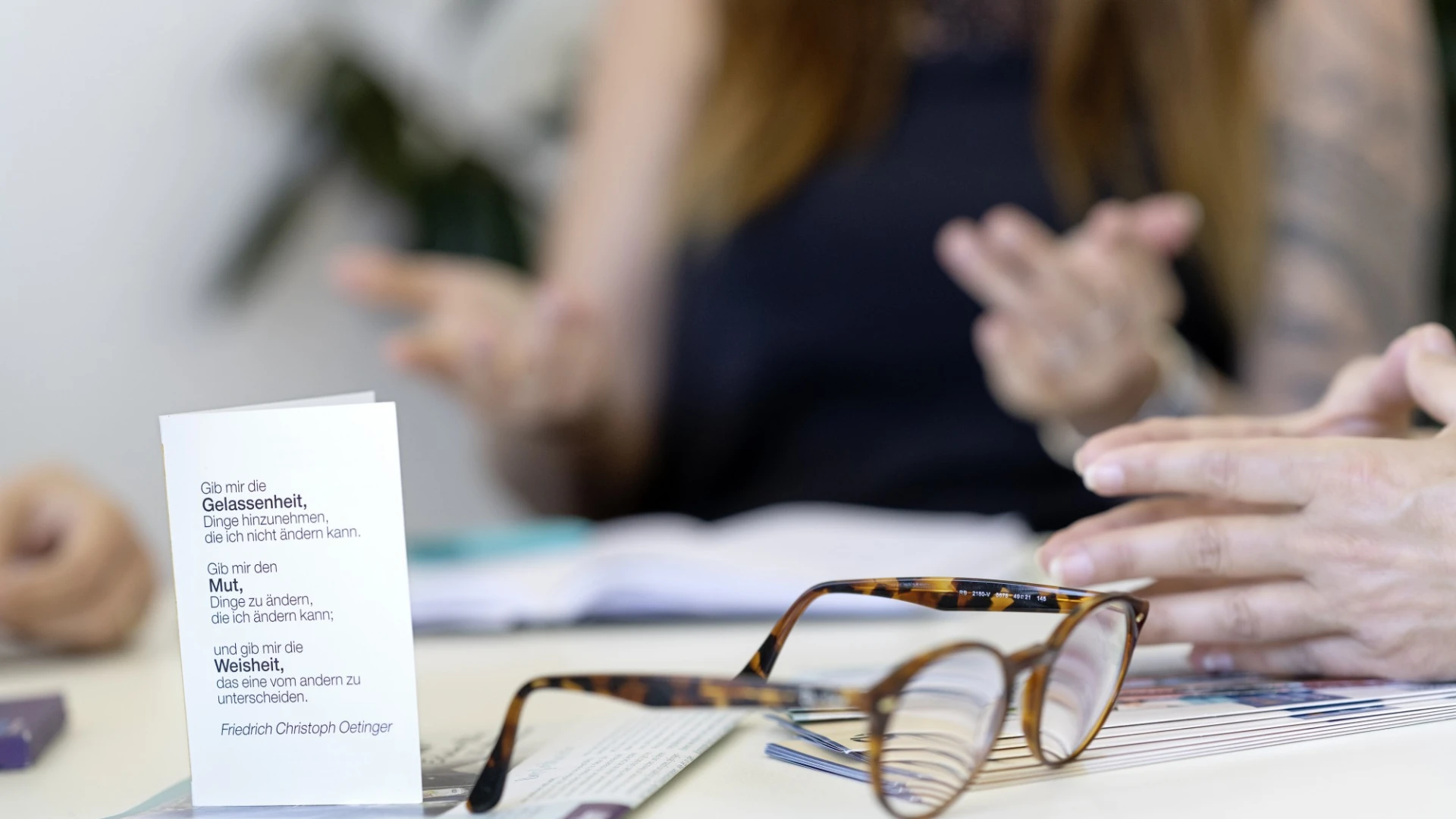 Braune Lesebrille liegt auf einem Tisch neben einem wei&szlig;en Dokument, w&auml;hrend im Hintergrund verschwommen eine Person sitzt und Handgesten macht.
