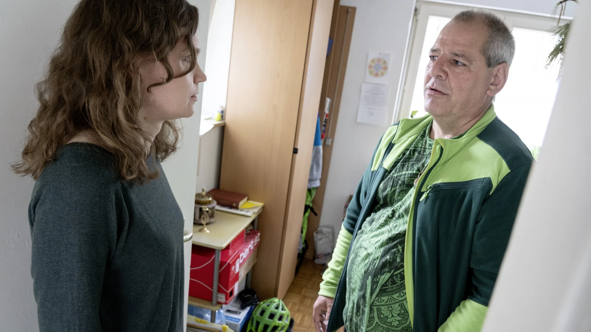 Eine Mitarbeiterin und ein Mann stehen sich in einem Raum gegen&uuml;ber, die Frau in einem grauen Pullover und der Mann in einer gr&uuml;n-schwarzen Jacke. Im Hintergrund ist ein Schrank und ein Fahrradhelm zu sehen.