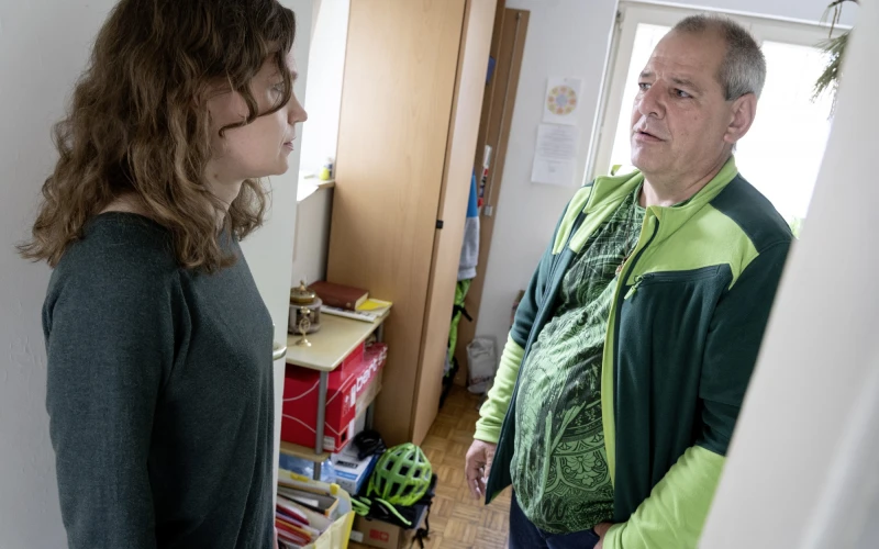 Eine Mitarbeiterin und ein Mann stehen sich in einem Raum gegen&uuml;ber, die Frau in einem grauen Pullover und der Mann in einer gr&uuml;n-schwarzen Jacke. Im Hintergrund ist ein Schrank und ein Fahrradhelm zu sehen.