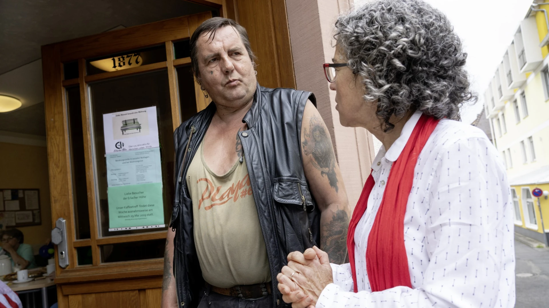Ein Mann mit Lederweste und Tattoo steht vor einem Holzt&uuml;r und unterh&auml;lt sich mit einer Frau mit grauen, lockigen Haaren in wei&szlig;er Bluse und einem roten Schal.