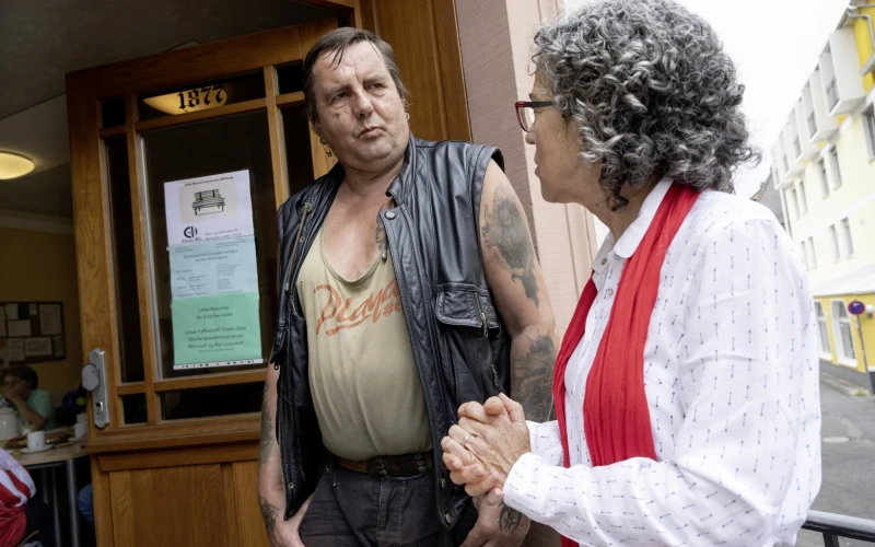 Ein Mann mit Lederweste und Tattoo steht vor einem Holzt&uuml;r und unterh&auml;lt sich mit einer Frau mit grauen, lockigen Haaren in wei&szlig;er Bluse und einem roten Schal.