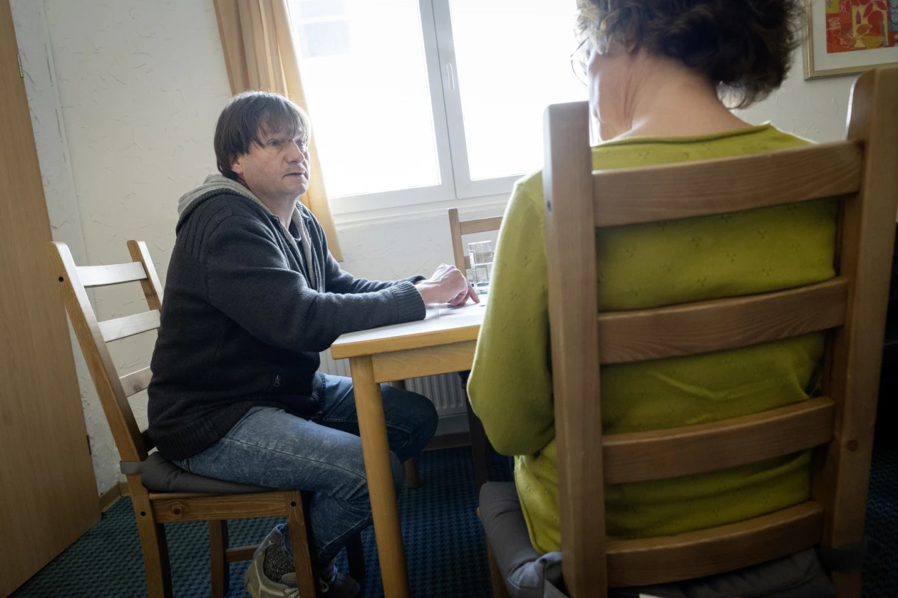 Ein Mann in einem grauen Pullover sitzt an einem Holztisch und spricht mit einer Mitarbeiterin in einem gr&uuml;nen Oberteil, deren R&uuml;cken zur Kamera gerichtet ist. Sie sitzen in einem hellen Raum mit Fenster und Holzst&uuml;hlen.