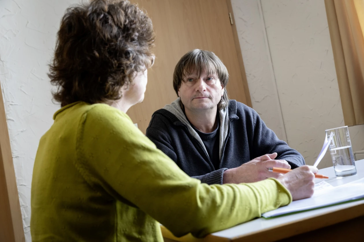 Eine Mitarbeiterin und ein Mann sitzen an einem Tisch. Die Mitarbeiterin tr&auml;gt einen gr&uuml;nen Pullover und ist von hinten zu sehen, der Mann in einem dunklen Pullover sitzt ihr gegen&uuml;ber. Ein Wasserglas steht zwischen ihnen auf dem Tisch.