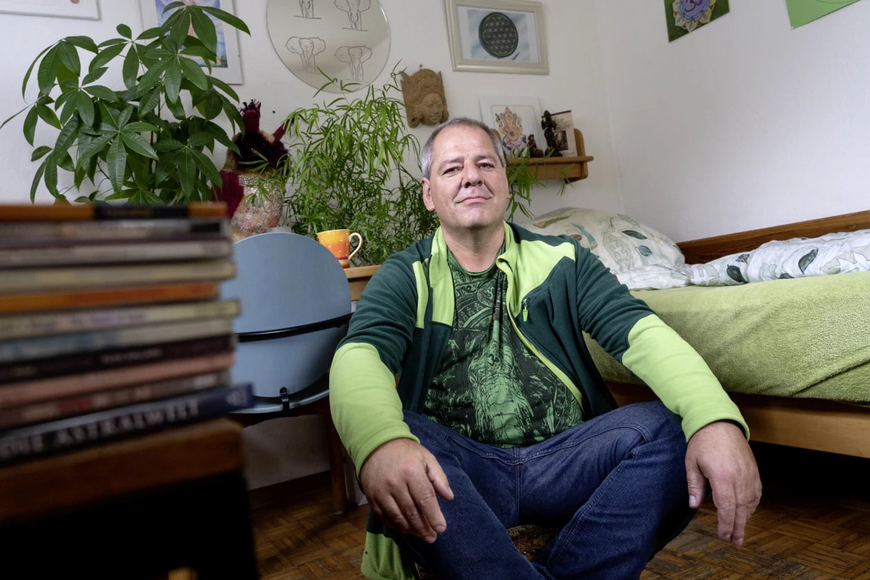 Ein l&auml;chelnder Mann in gr&uuml;n-blauer Jacke sitzt vor einem Bett in einem Zimmer. Im Hintergrund sind Zimmerpflanzen, B&uuml;cherstapel und Mobiliar.