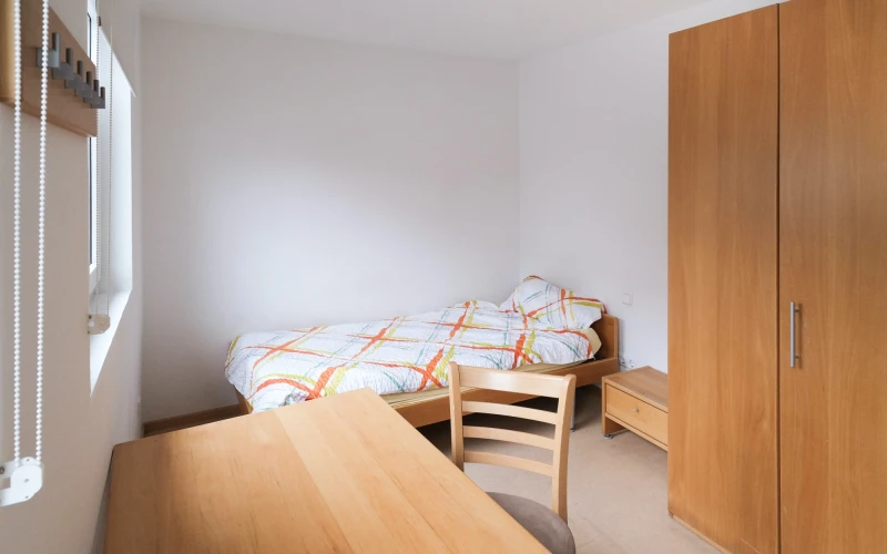 Einzelzimmer mit Holzm&ouml;beln, bestehend aus einem Bett, einem Holzschrank und einem Holztisch mit Stuhl. Das Zimmer hat wei&szlig;e W&auml;nde und eine Jalousie am Fenster, die Bettw&auml;sche ist farbig bezogen.