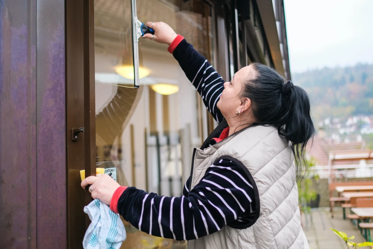 Eine Frau in einem gestreiften Pullover und einer grauen Weste reinigt ein Fenster.