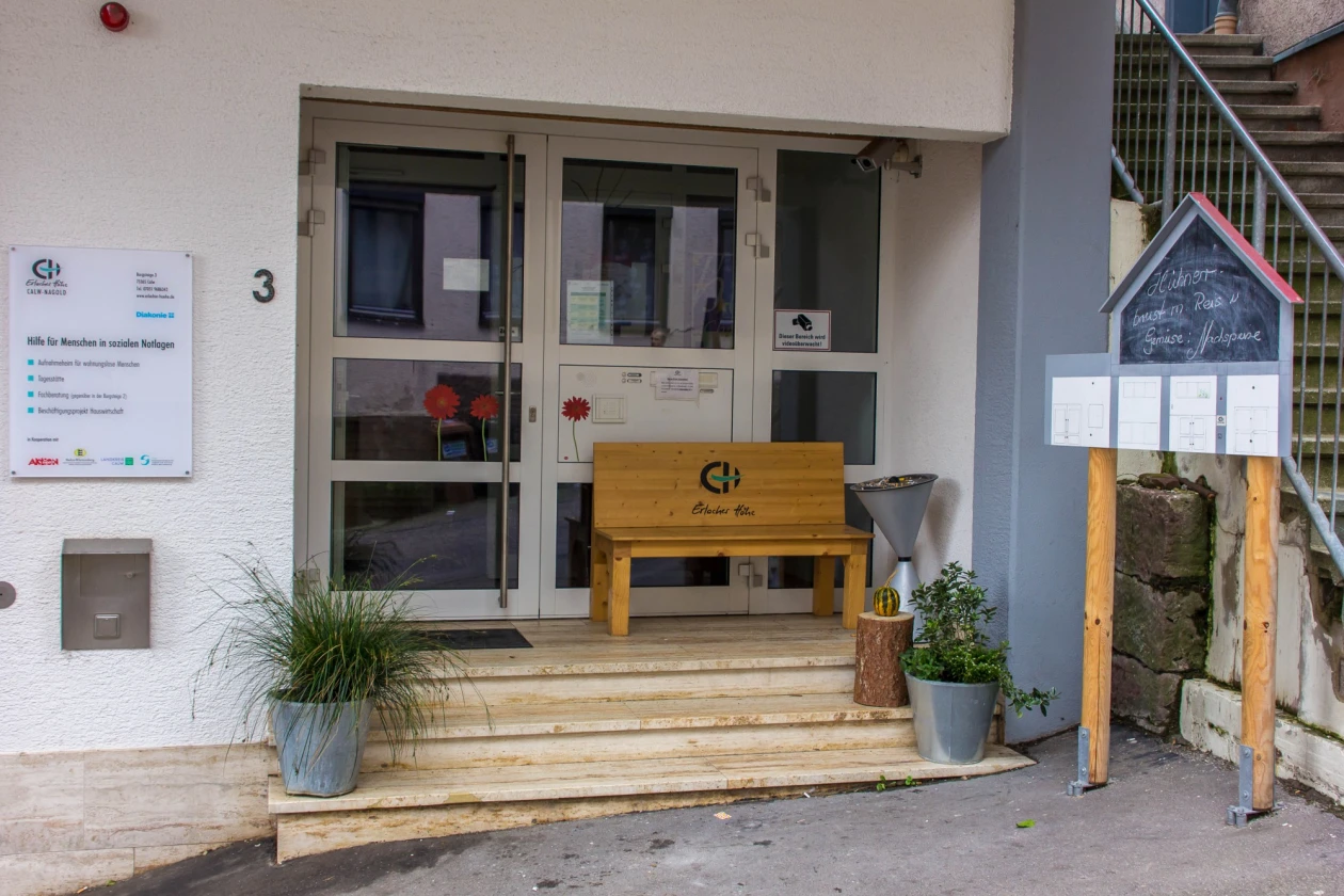 Holzbank mit EH Logo vor einer Glaseingangst&uuml;re mit Treppe mit Blument&ouml;pfen und einer grauen Infotafel. Auf dem Hinweisschild an der Wand steht "Hilfe f&uuml;r Menschen in sozialen Notlagen.