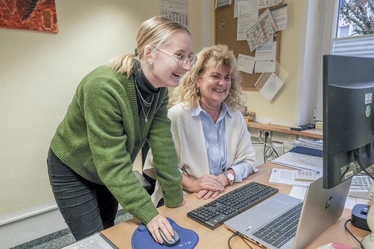 Zwei lachende Frauen arbeiten gemeinsam an einem Computer am Schreibtisch. Eine Frau in einem gr&uuml;nen Pullover und einer Brille lehnt sich vor und bedient die Maus, w&auml;hrend die andere Frau in einer wei&szlig;en hellblauen Bluse mit wei&szlig;er Jacke daneben sitzt.