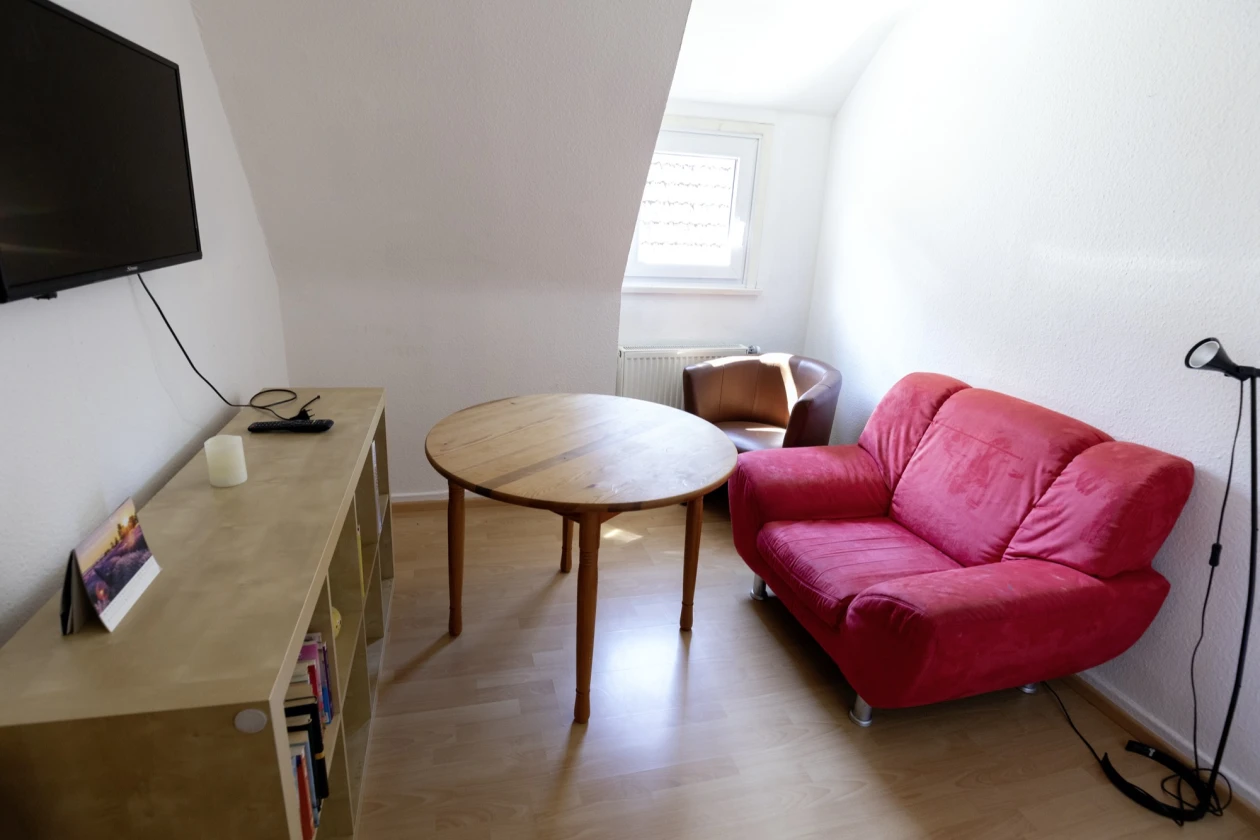 Kleiner Dachraum mit minimalistischer Einrichtung: Ein rosaroter Sessel, ein runder Holztisch, ein Sofa und ein langes Sideboard mit einer Fernseh-Ecke. Das Zimmer hat ein Fenster und helle, holzfarbene B&ouml;den.