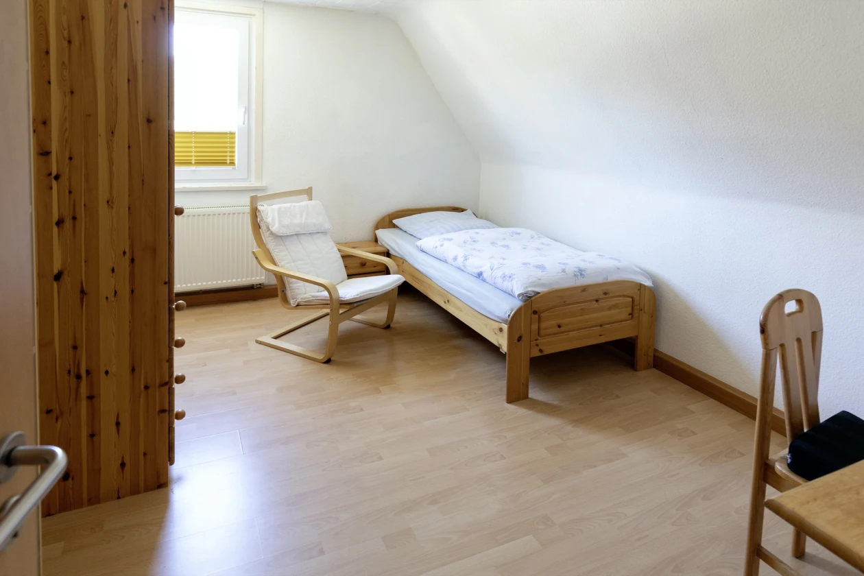Einfaches Einzelzimmer mit Holzbett, wei&szlig;en W&auml;nden und Parkettboden. Ein Holzsessel steht neben dem Bett, im Vordergrund steht ein Holztisch mit Stuhl. Durch das Fenster f&auml;llt nat&uuml;rliches Licht in den Raum.