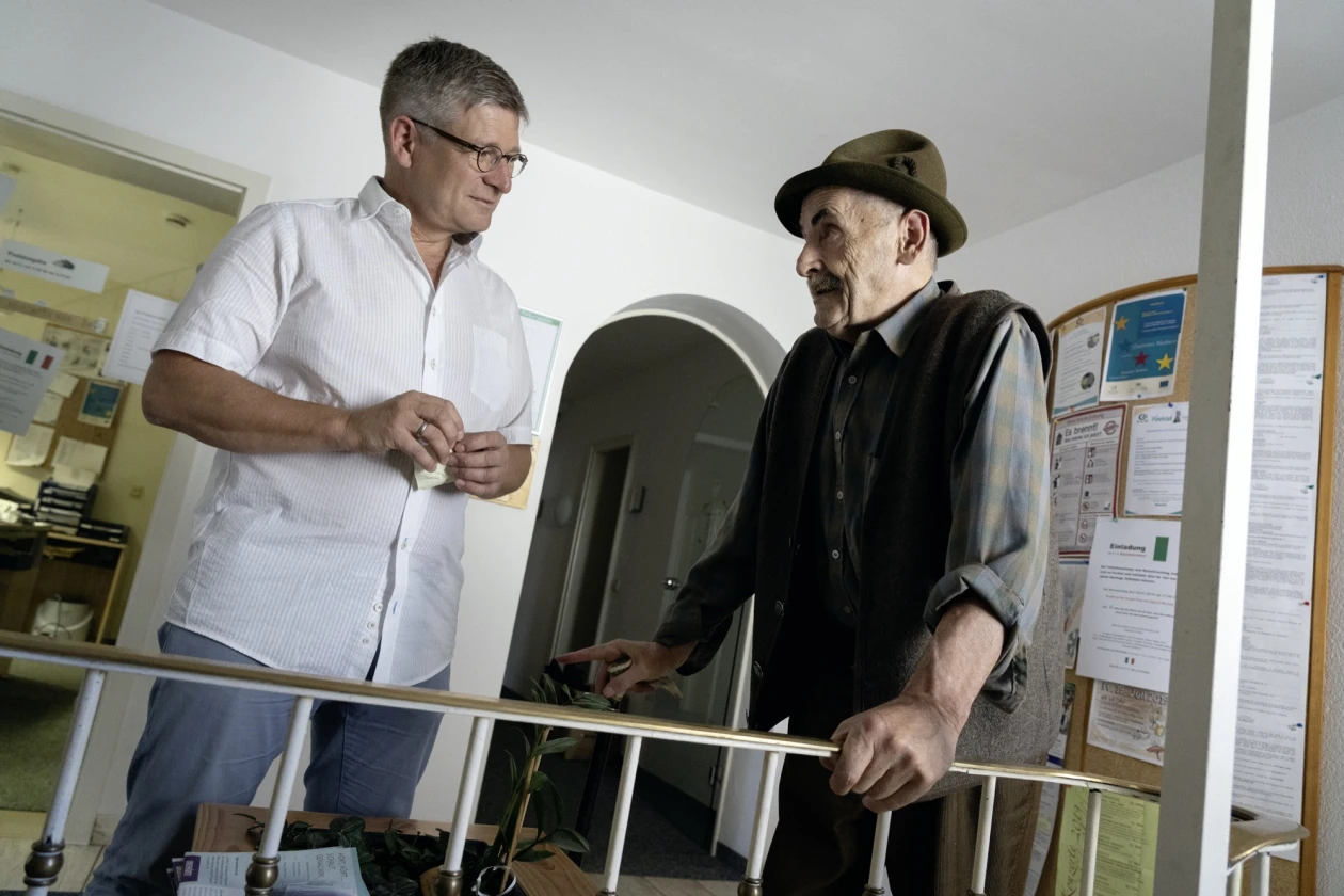 Ein Mitarbeiter mit wei&szlig;em Hemd ber&auml;t einen &auml;lteren Mann in M&uuml;tze in einer Einrichtung, w&auml;hrend beide an einem Gel&auml;nder stehen.