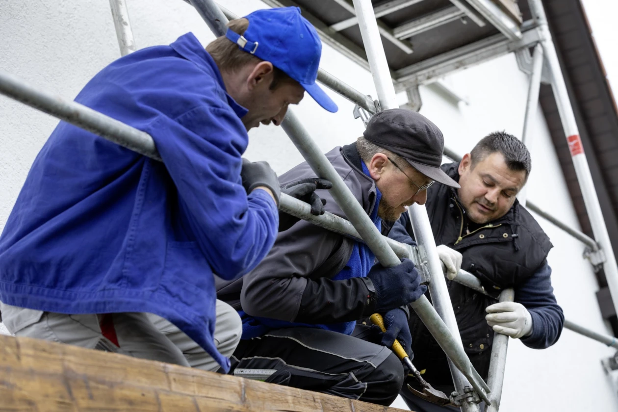 Drei Arbeiter in Blau- und Schwarzt&ouml;nen arbeiten am Bauger&uuml;st. Der Mann in der Mitte hat einen Hammer in der Hand.