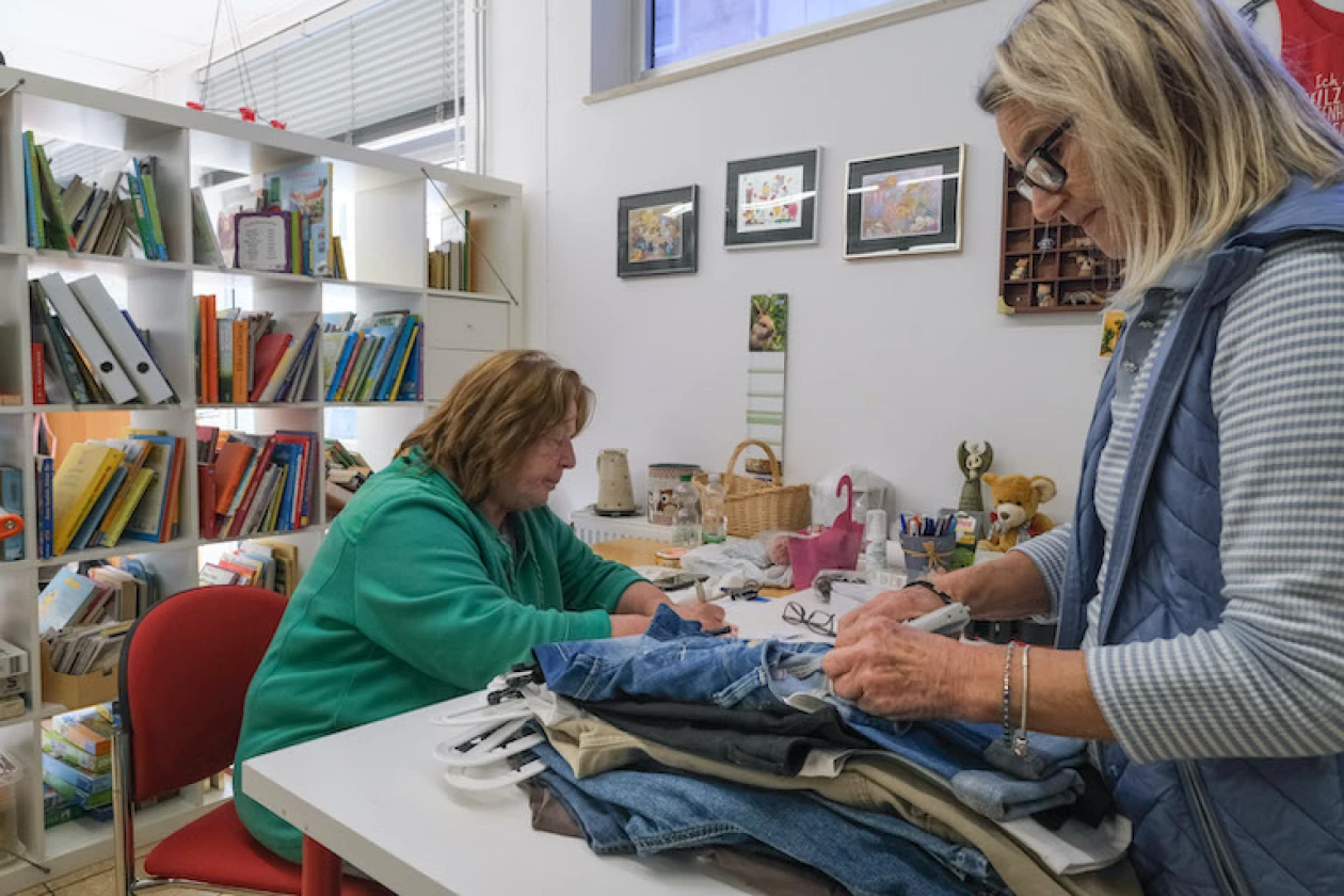 Zwei Frauen arbeiten an einem Tisch mit Kleidungsst&uuml;cken. Im Hintergrund befindet sich ein Regal mit B&uuml;chern und Ordnern.