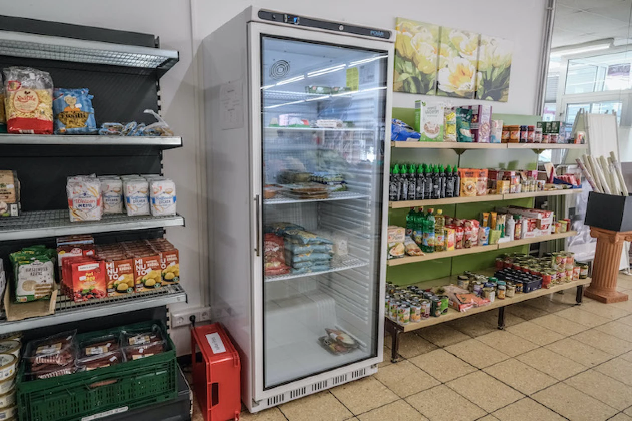 Gro&szlig;er K&uuml;hlschrank mit Glast&uuml;r in einem Lebensmittelgesch&auml;ft, umgeben von Regalen gef&uuml;llt mit verschiedenen Produkten und Lebensmitteln.