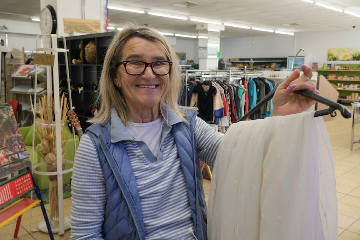 L&auml;chelnde Frau mit Brille in einem gestreiften Shirt und einer violetten Weste, die ein wei&szlig;en Kleidungsst&uuml;ck auf einem Kleiderb&uuml;gel zeigt.