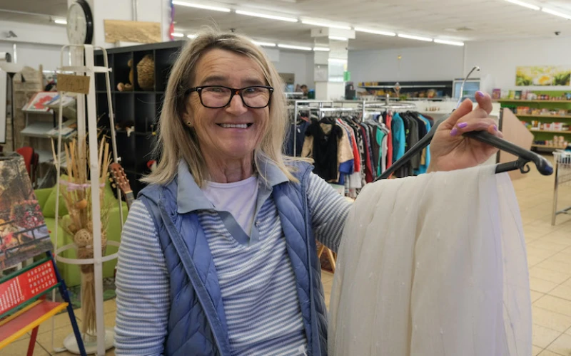 L&auml;chelnde Frau mit Brille in einem gestreiften Shirt und einer violetten Weste, die ein wei&szlig;en Kleidungsst&uuml;ck auf einem Kleiderb&uuml;gel zeigt.