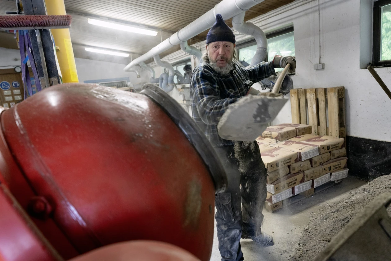 Arbeiter in karierter Jacke und M&uuml;tze bef&uuml;llt einen roten Betonmischer. Im Hintergrund sind Holzpaletten mit Zements&auml;cken.