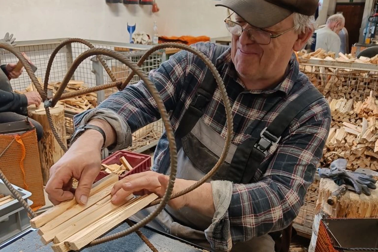 Ein Mann in kariertem Hemd und M&uuml;tze f&uuml;llt das herzf&ouml;rmige Metallgestell mit Anz&uuml;ndholz. im Hintergrund sieht man weiteres Anz&uuml;ndholz.