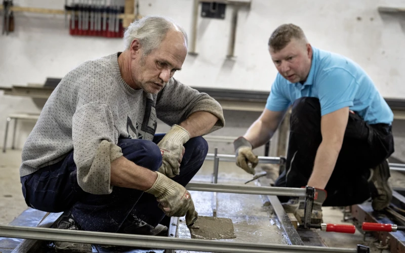 Zwei M&auml;nner bearbeiten ein Betonwerkst&uuml;ck in Schalung. Der j&uuml;ngere Mann tr&auml;gt ein hellblaues T-Shirt, der &auml;ltere einen grauen Pullover. Beide tragen Arbeitshandschuhe und konzentrieren sich auf ihre Arbeit.