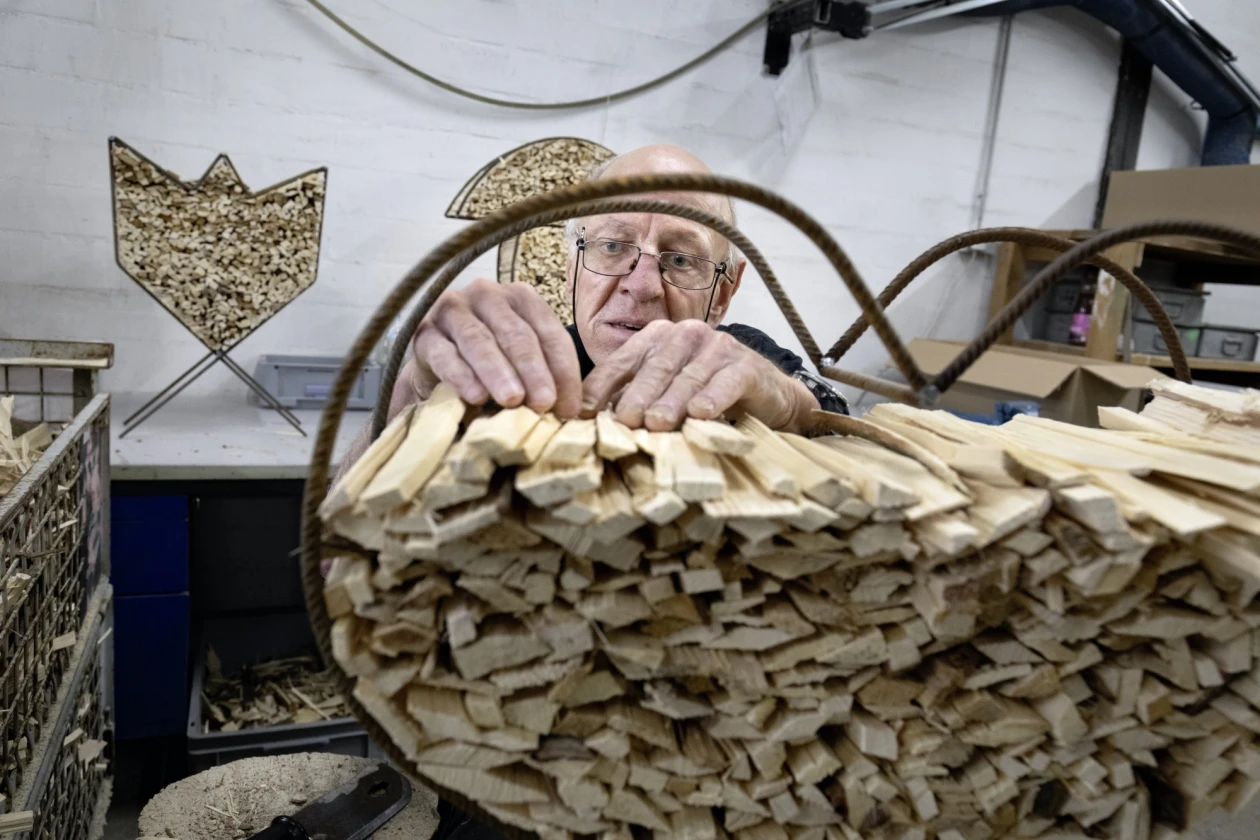 Ein Mann mit Brille sitzt und f&uuml;llt ein herzf&ouml;rmiges Metallgestell mit Anz&uuml;ndholz. Im Hintergrund sieht man weitere Metallgestelle, gef&uuml;llt mit Anz&uuml;ndholz.