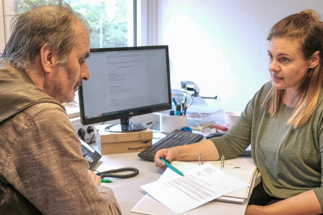 Ein Mann und eine Frau sitzen an einem Schreibtisch vor einem Computer. Auf einem Ordner liegt ein Dokument, das zu dem Mann gerichtet ist. Die Frau zeigt mit einem Kugelschreiber auf eine Textstelle.