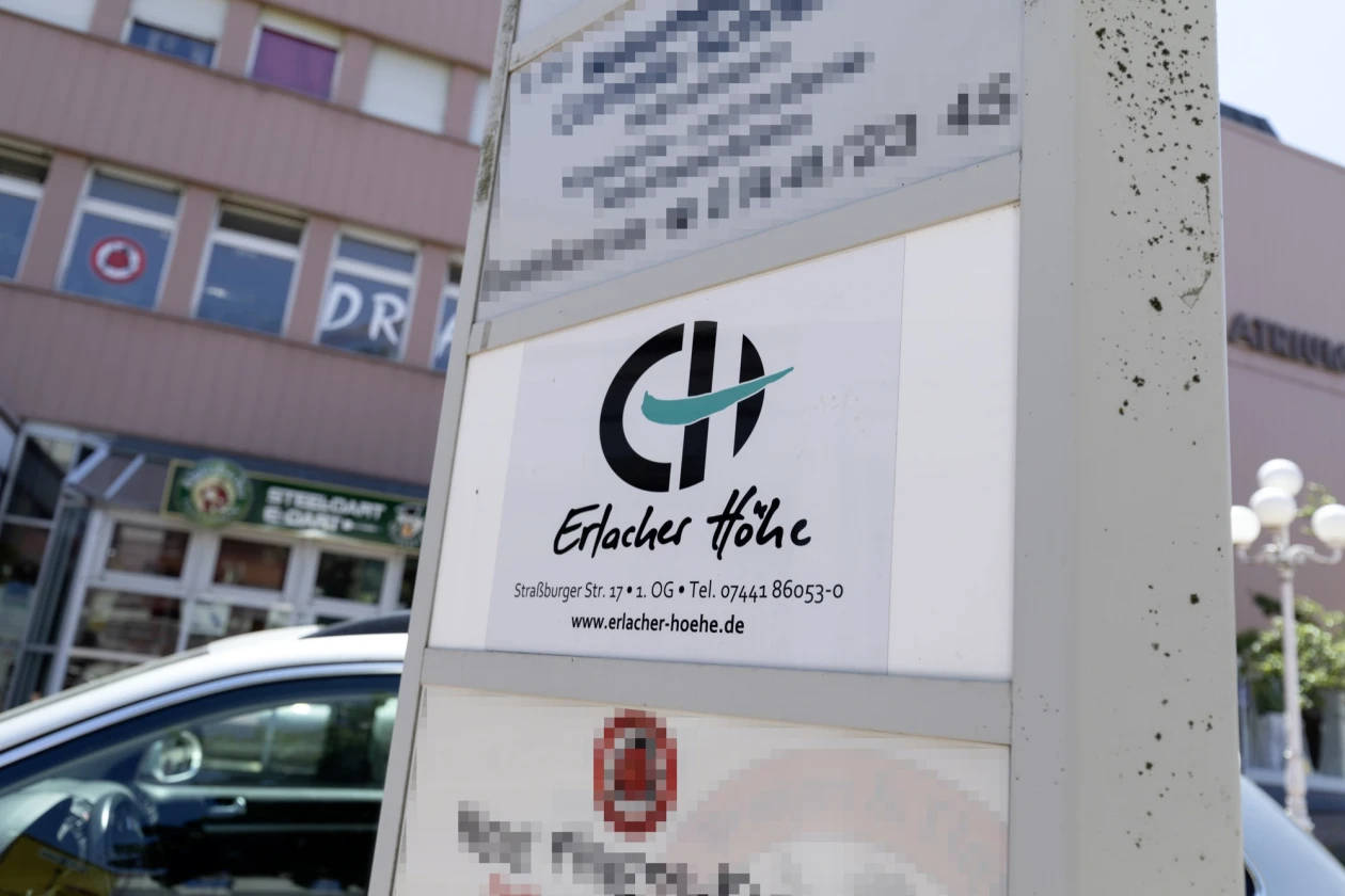 Schild mit dem Logo der Erlacher H&ouml;he. Im Hintergrund ist eine Geb&auml;ude an einer Stra&szlig;e zu sehen.