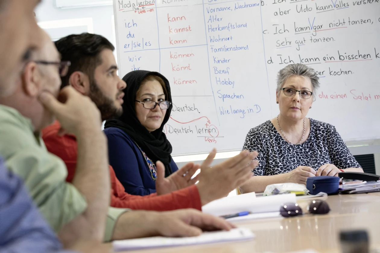 Vier Personen sitzen an einem Tisch, ein Mann gestikuliert, w&auml;hrend die anderen h&ouml;ren zu. Im Hintergrund ist ein Whiteboard zu sehen.