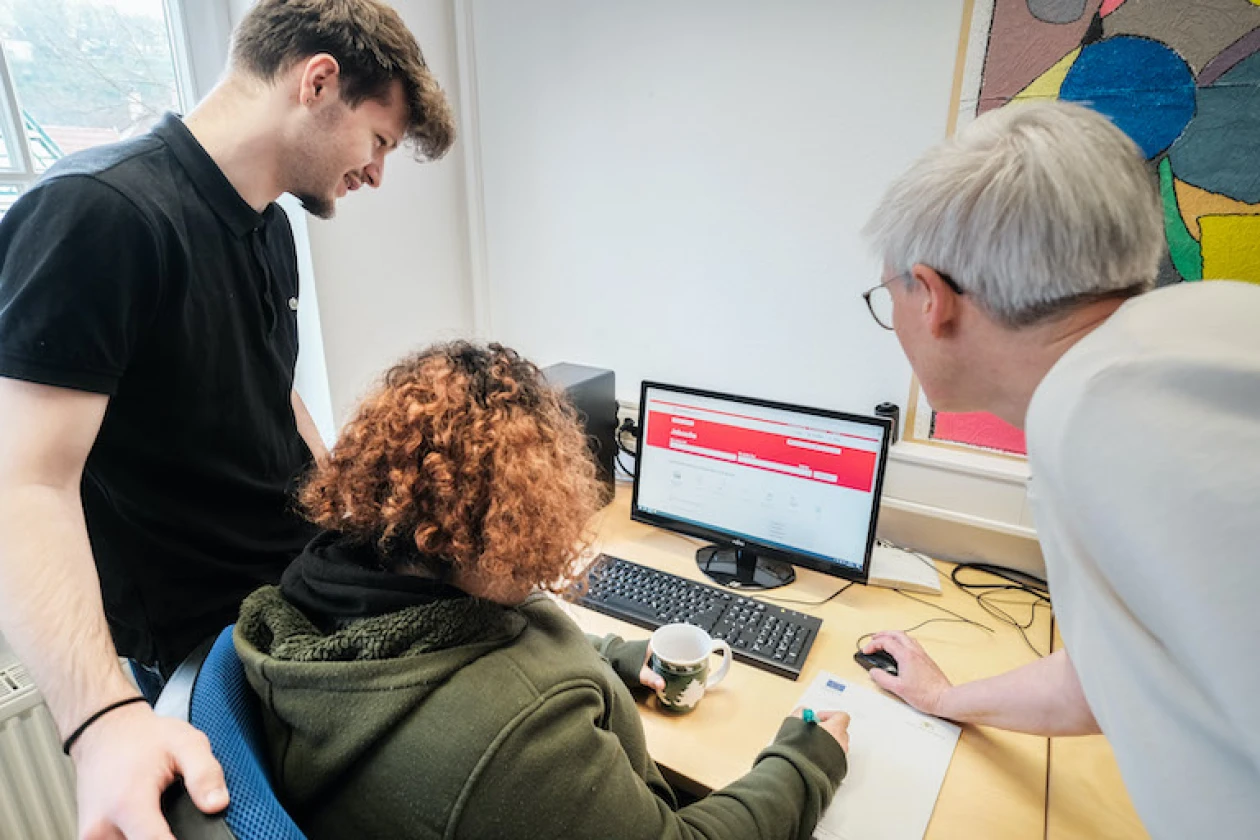 Zwei Personen stehen an einem Computerarbeitsplatz, w&auml;hrend eine Person im gr&uuml;nen Kapuzenpullover dort sitzt.