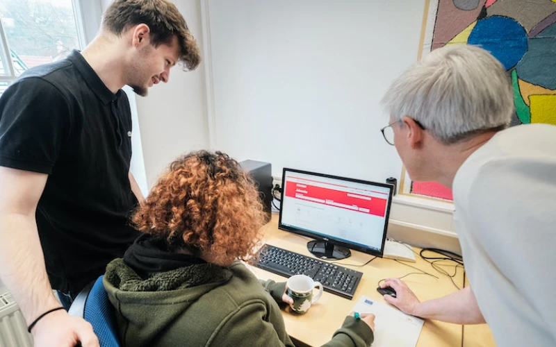 Zwei Personen stehen an einem Computerarbeitsplatz, w&auml;hrend eine Person im gr&uuml;nen Kapuzenpullover dort sitzt.