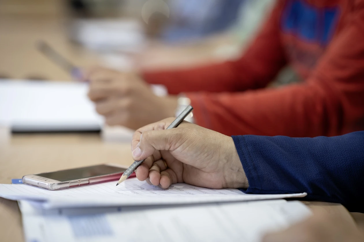 Zu sehen ist eine Hand mit Bleistift beim Schreiben auf Papier, daneben liegt ein Smartphone, im Hintergrund ist eine weitere Hand am Tisch zu sehen. Die Situation &auml;hnelt einer Seminarteilnahme.