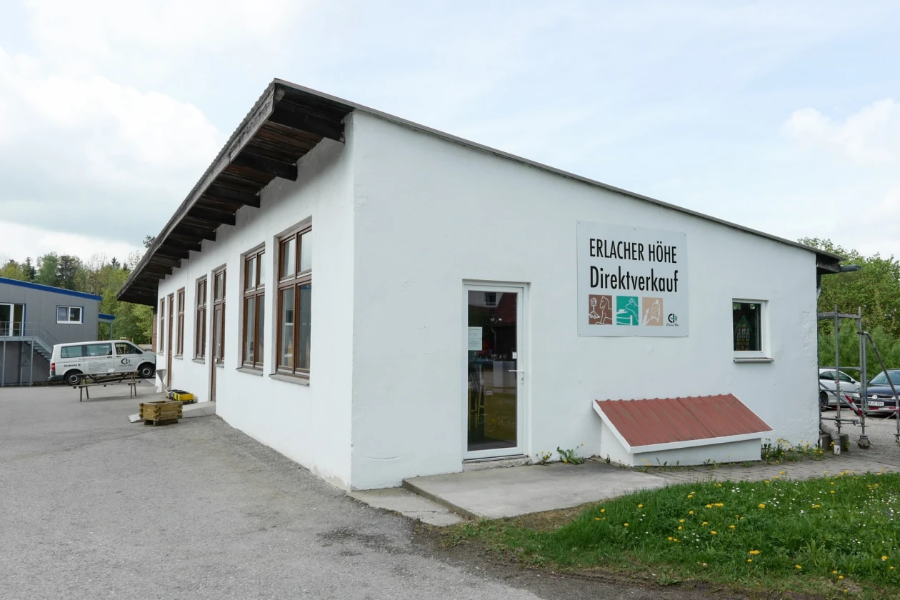 Wei&szlig;es Geb&auml;ude mit flachem Dach und gro&szlig;en Fenstern,  An der Wand ist ein Schild mit der Aufschrift "Erlacher H&ouml;he Direktverkauf". Im Hintergrund sind Parkpl&auml;tze mit parkenden Autos zu sehen.