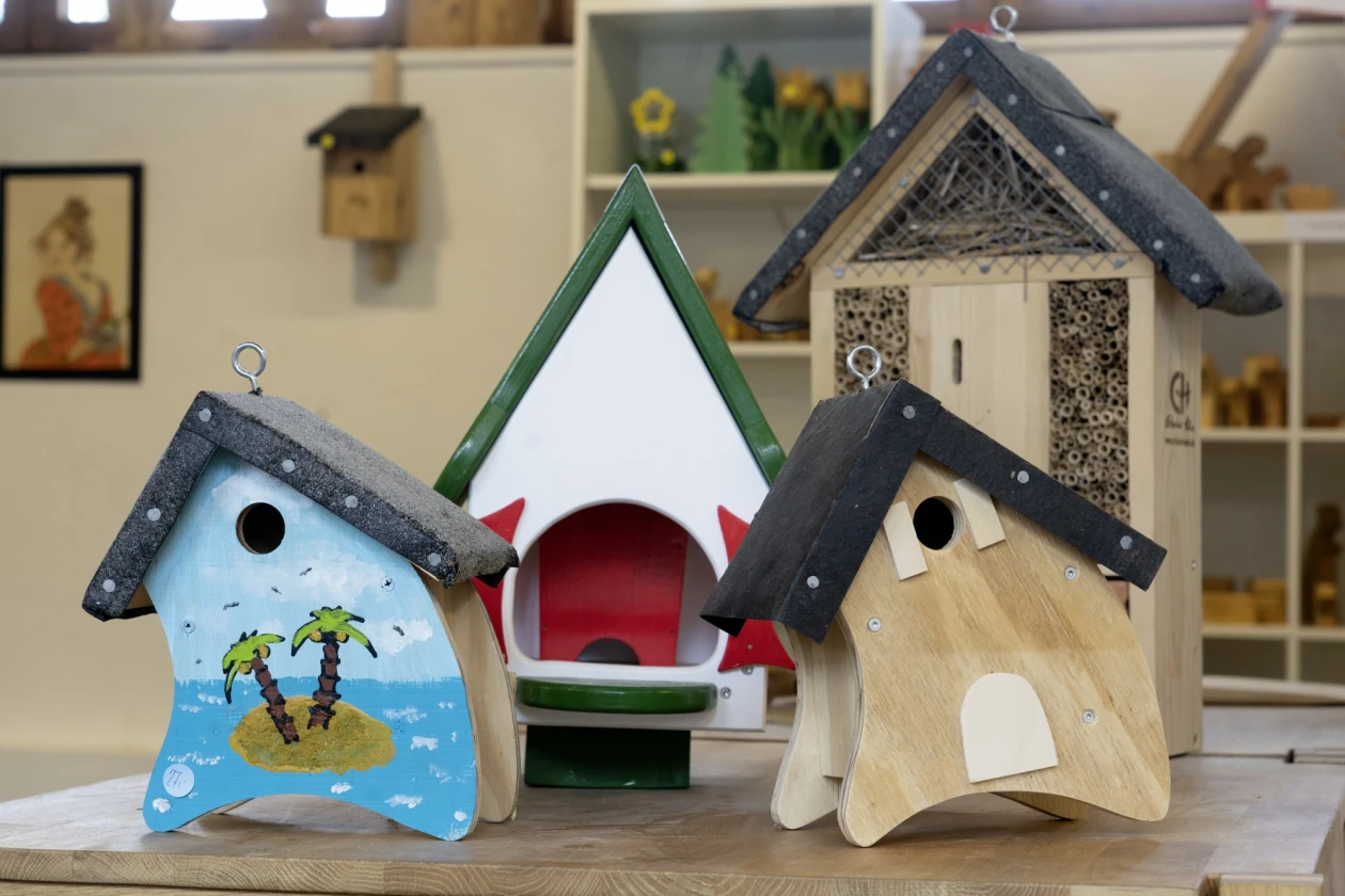 Vier dekorativ gestaltete Vogelh&auml;user in verschiedenen Farben und Designs, darunter ein blau-wei&szlig;es mit Palmen-Motiv, ein wei&szlig;-rotes, ein natur-farbenes und ein weiteres grau-wei&szlig;es Modell, arrangiert auf einer Holzoberfl&auml;che mit unscharfem Hintergrund.