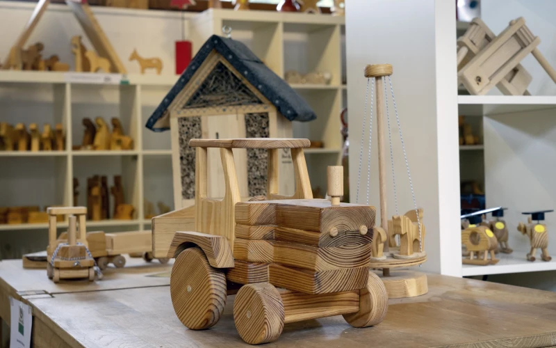 Handgefertigter Spielzeug-Traktor aus Holz mit Details wie R&auml;dern und Kabine auf einem Tisch. Im Hintergrund sieht man weitere handgefertigte Holzspielzeuge.