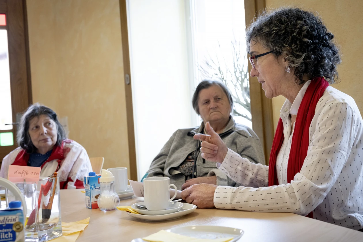 Drei Frauen sitzen an einem Tisch mit Kaffeegedecken. Die Frau im wei&szlig;en Pullover und rotem Schal erz&auml;hlt, w&auml;hrend die anderen beiden aufmerksam zuh&ouml;ren.