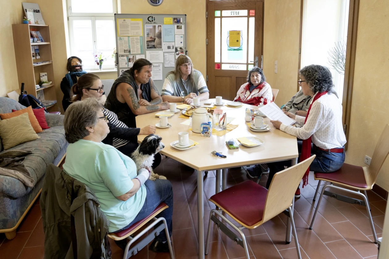 Mehrere Personen sitzen an einem sechseckigen Tisch in einem Gemeinschaftsraum mit Kaffee und Geb&auml;ck. Im Raum befinden sich Pinnwand, Regale, ein Sofa mit Kissen und Fenster.