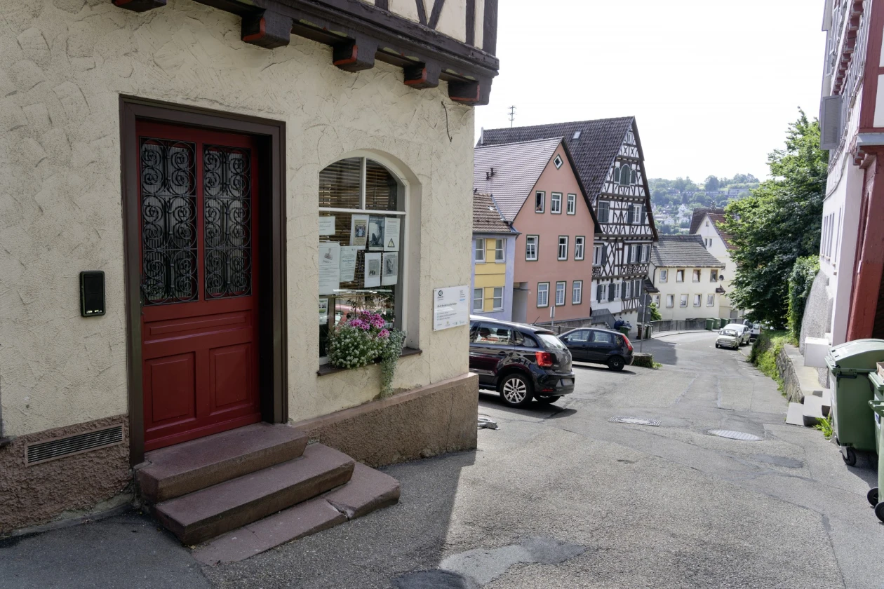 Eine Stra&szlig;e mit historischen Fachwerkh&auml;usern und geparkten Autos. Das Geb&auml;ude im Vordergrund hat eine rote Eingangst&uuml;r, eine Steintreppe und Blumen auf der Fensterbank.