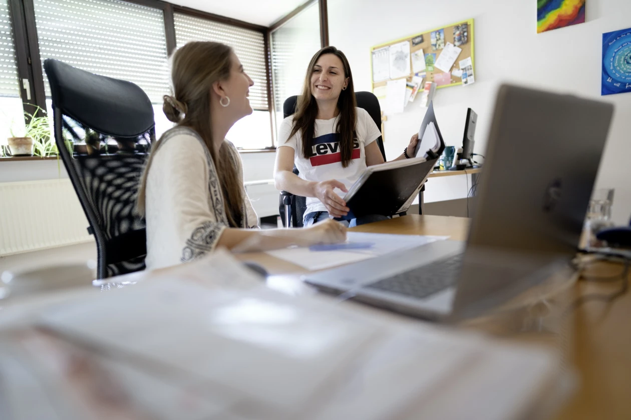 Zwei Frauen sitzen an einem Schreibtisch und unterhalten sich l&auml;chelnd. Laptops, eine Pinnwand und B&uuml;romaterialien sind im Hintergrund zu sehen.