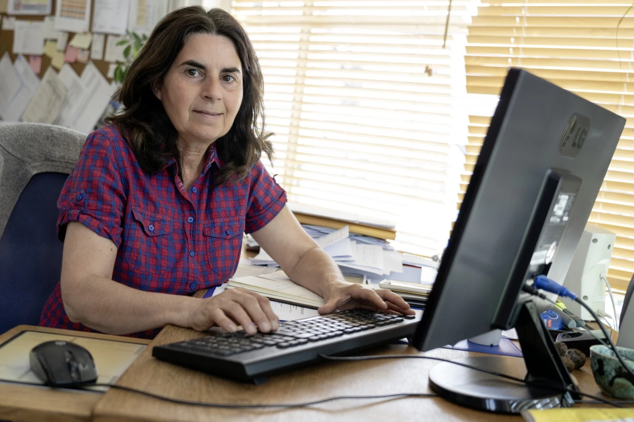 Eine Frau mit dunklen langen Haaren in rot-kariertem Hemd sitzt am Schreibtisch und arbeitet am Computer. Sie blickt direkt in die Kamera. Tastatur und Monitor stehen im Vordergrund, im Hintergrund sind am Fenster Jalousien zu sehen.