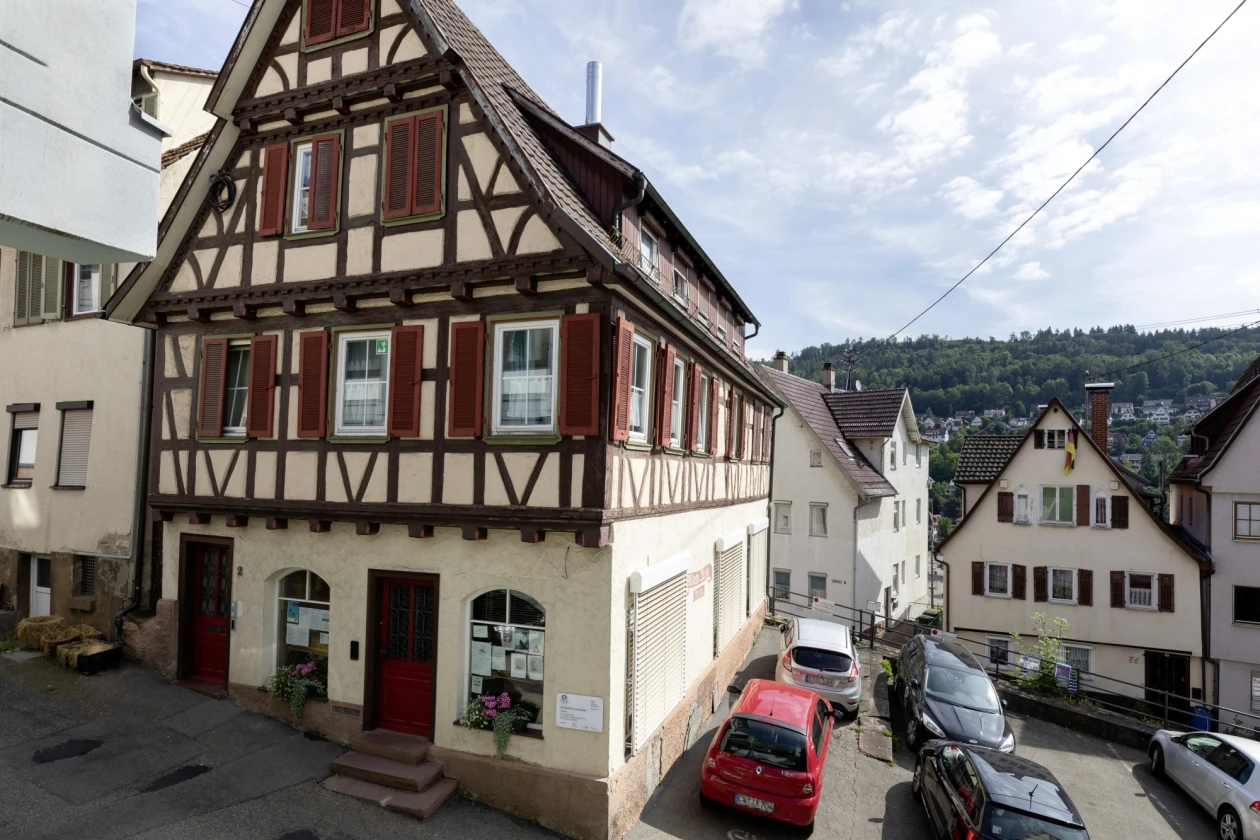 Fachwerkhaus in Hanglage an einer engen Stra&szlig;e mit parkenden Autos. Im Hintergrund sind weitere Geb&auml;ude eines Stadtkerns in unterschiedlichen Baustilen und Fassadenfarben zu sehen.