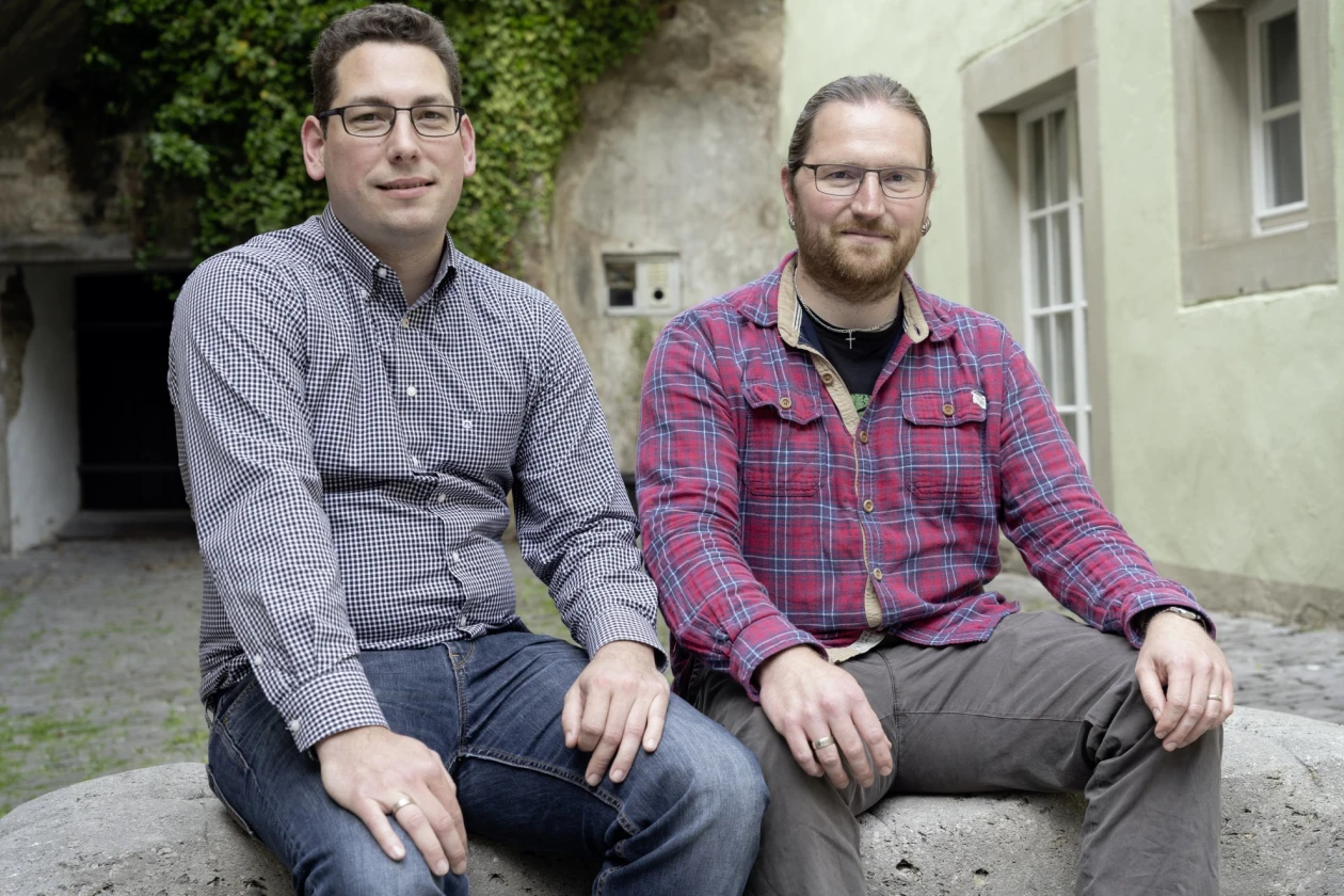Zwei M&auml;nner sitzen nebeneinander vor einem Geb&auml;ude. Der linke tr&auml;gt ein kleinkariertes Hemd und eine Brille, der rechte tr&auml;gt ein rotes Karohemd und tr&auml;gt ebenfalls eine Brille. Sie blicken l&auml;chelnd in die Kamera.