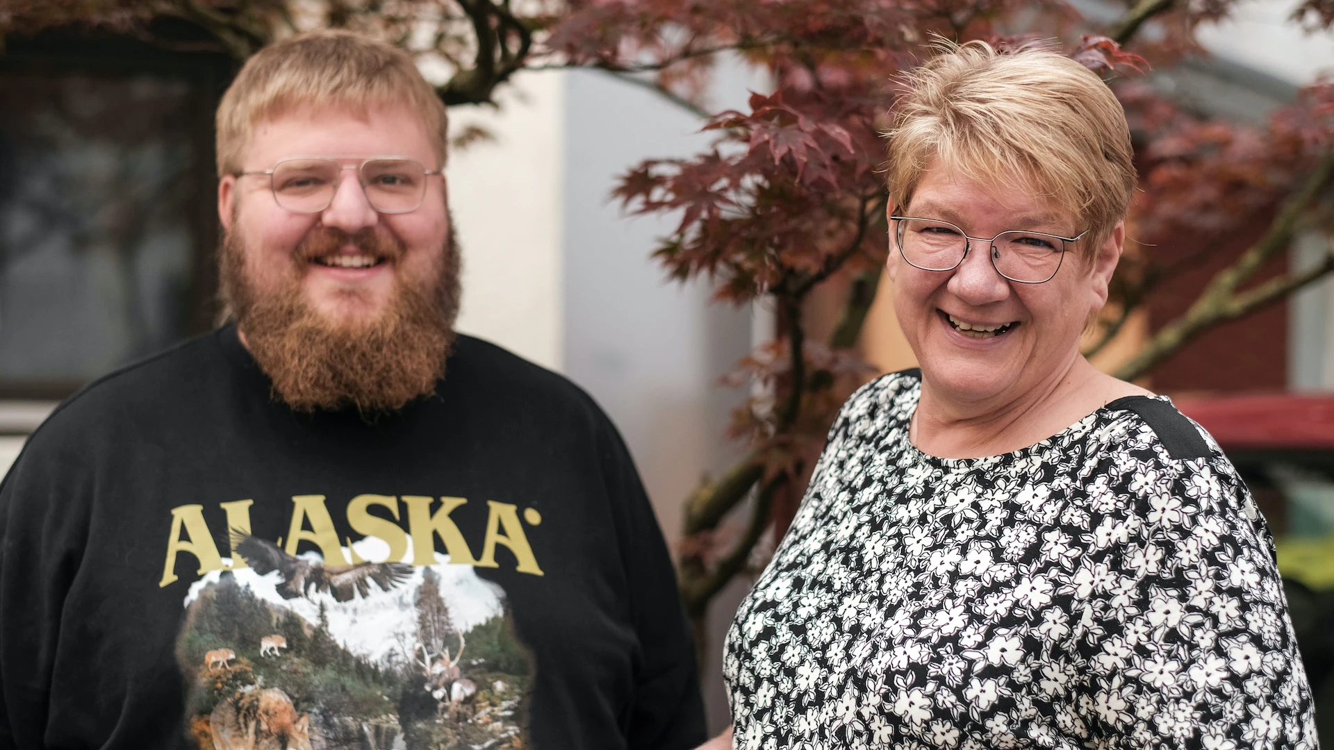 Ein Mann und eine Frau l&auml;cheln in die Kamera. Der blondhaarige Mann mit Bart tr&auml;gt ein dunkles T-Shirt an mit der Aufschrift "Alaska". Die blondhaarige Frau tr&auml;gt ein schwarz-wei&szlig; gemustertes Oberteil. Beide stehen vor einem Baum mit r&ouml;tlichen Bl&auml;ttern.