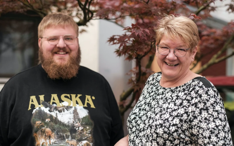 Ein Mann und eine Frau l&auml;cheln in die Kamera. Der blondhaarige Mann mit Bart tr&auml;gt ein dunkles T-Shirt an mit der Aufschrift "Alaska". Die blondhaarige Frau tr&auml;gt ein schwarz-wei&szlig; gemustertes Oberteil. Beide stehen vor einem Baum mit r&ouml;tlichen Bl&auml;ttern.