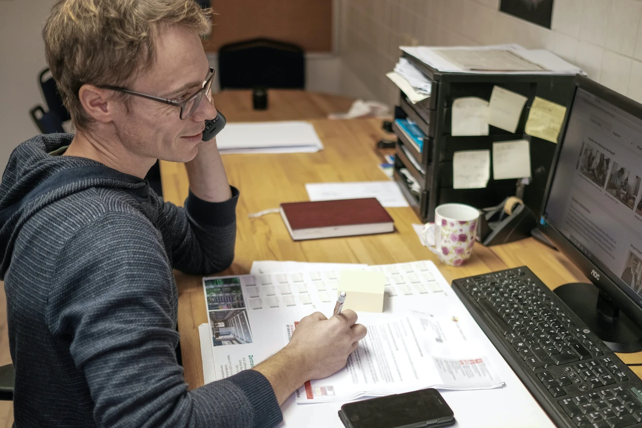Ein Mann mit Brille und dunklem Kapuzenpulli sitzt an einem Schreibtisch. Er h&auml;lt das Telefon am linken Ohr und blickt dabei auf den Bildschirm. Auf dem Schreibtisch sind Dokumente, ein Handy, eine Tasse sowie weiteres B&uuml;romaterial zu sehen.