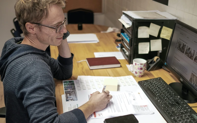 Ein Mann mit Brille und dunklem Kapuzenpulli sitzt an einem Schreibtisch. Er h&auml;lt das Telefon am linken Ohr und blickt dabei auf den Bildschirm. Auf dem Schreibtisch sind Dokumente, ein Handy, eine Tasse sowie weiteres B&uuml;romaterial zu sehen.