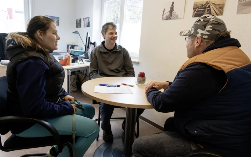 Drei Personen sitzen an einem runden Tisch in einem B&uuml;ro mit Fenster. Eine Frau und zwei M&auml;nner unterhalten sich miteinander, w&auml;hrend Papiere und Kulis auf dem Tisch liegen. Der Raum ist hell und mit Bildern an den W&auml;nden dekoriert.