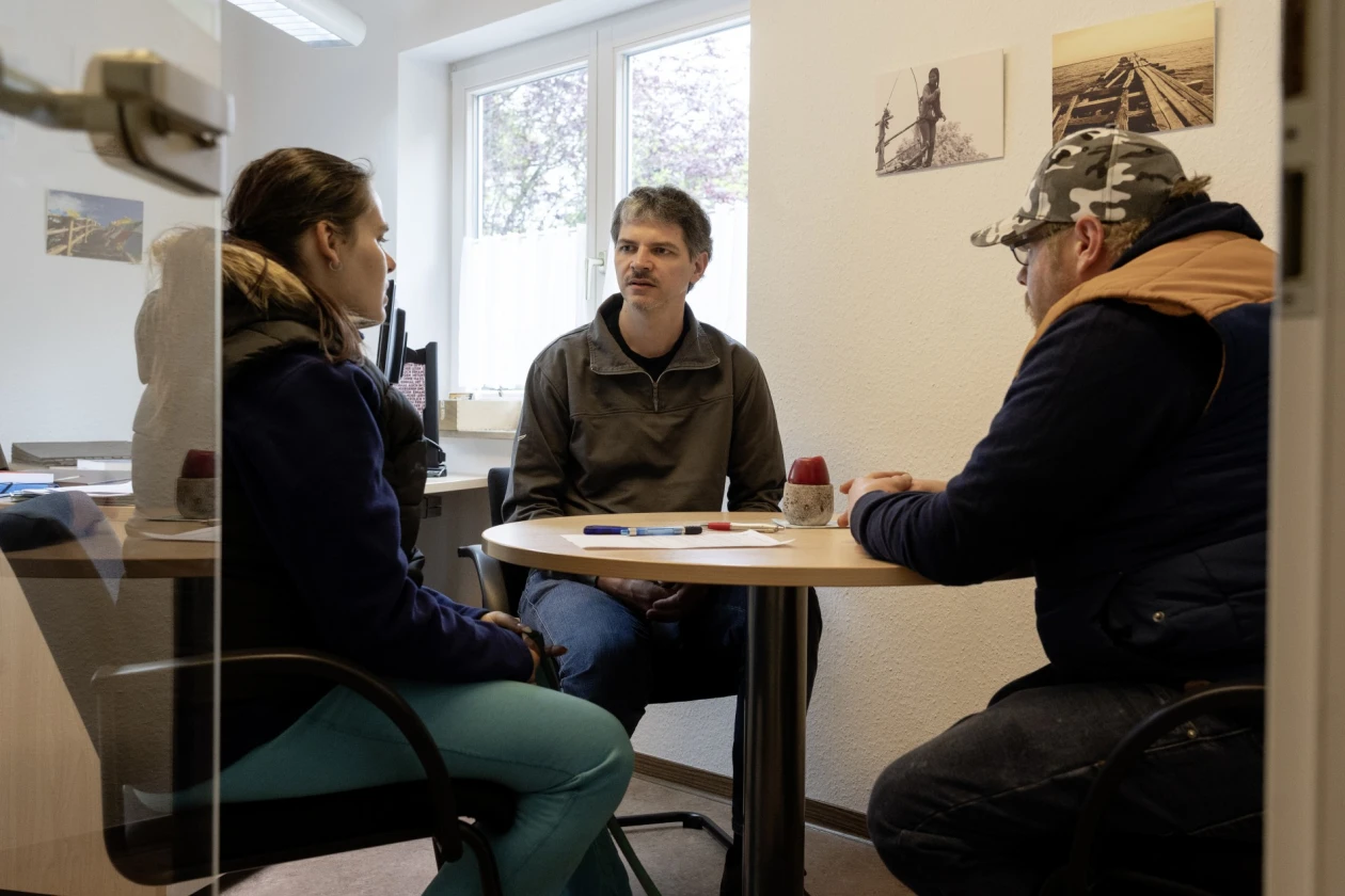 Drei Personen sitzen an einem runden Tisch in einem hellen B&uuml;roraum mit Fenster und f&uuml;hren ein Gespr&auml;ch. Auf dem Tisch liegen Dokumente. An den W&auml;nden h&auml;ngen Bilder.