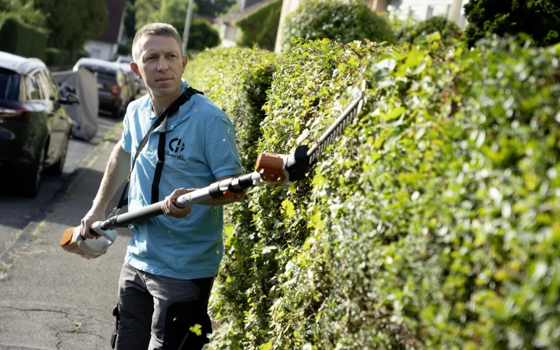 Ein Mann im blauen T-Shirt mit Logo der Erlacher H&ouml;he steht auf einem Gehweg und schneidet eine gr&uuml;ne Hecke mit einer Heckenschere. Im Hintergrund sind parkende Autos und H&auml;user zu sehen sind.