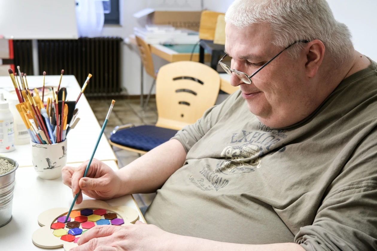 Ein Mann mit Brille sitzt an einem Tisch und h&auml;lt einen Pinsel in der Hand mit dem er bunte Pl&auml;ttchen in einer Holzblumen anordnet. Neben ihm stehen mehrere Pinsel in einem Becher und im Hintergrund ein weiterer Tisch mit St&uuml;hlen. Er tr&auml;gt ein graues T-Shirt und hat Freude an seiner kreativen Besch&auml;ftigung.