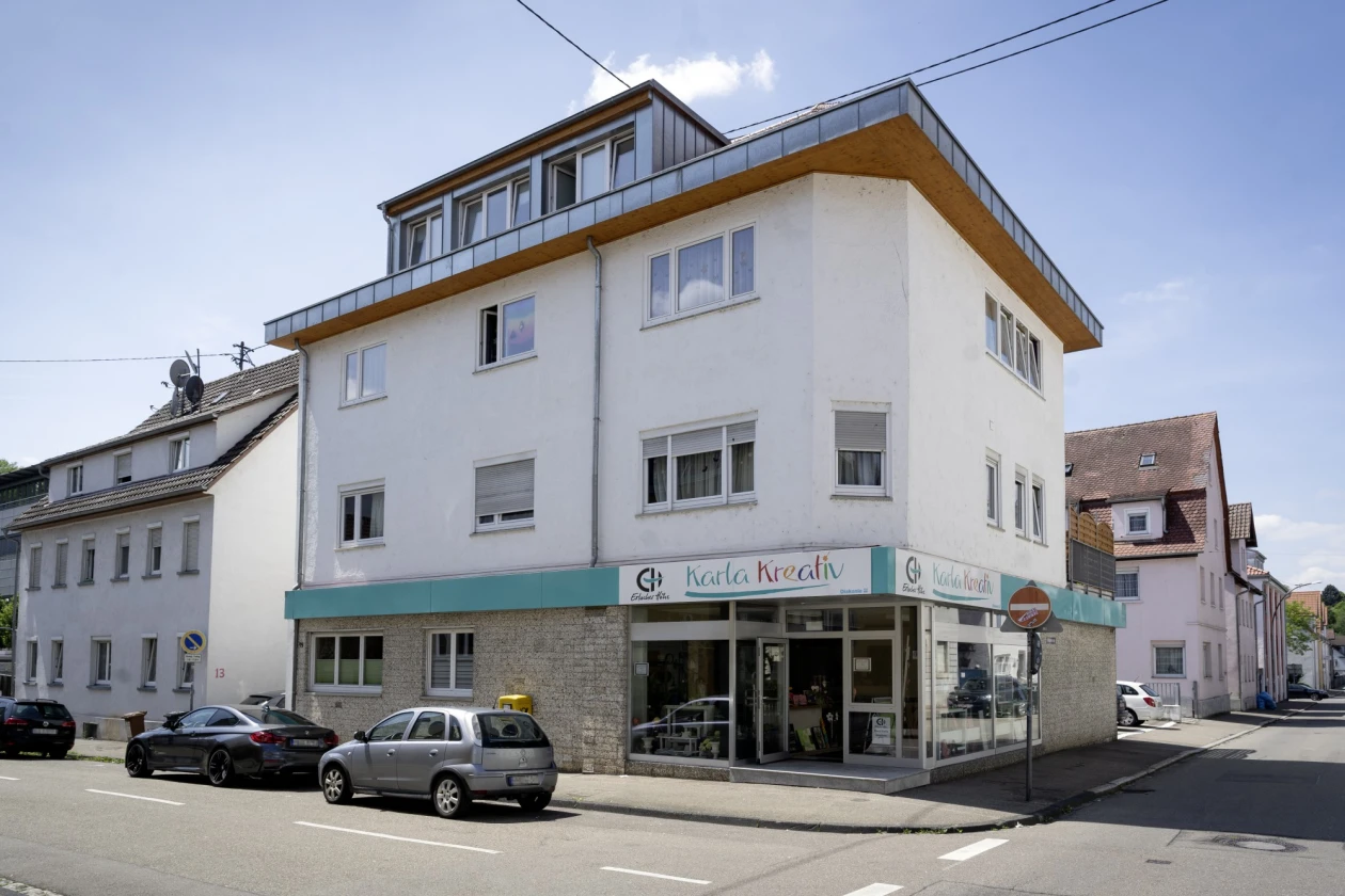 Ein wei&szlig;es Haus an einer Stra&szlig;enecke mit einem Ladenlokal im Erdgeschoss. Vor dem Haus ist das Schild &bdquo;Karla Kreativ&ldquo; mit dem Erlacher H&ouml;he-Logo zu sehen. Davor sind geparkte Autos, und im Hintergrund sind weitere H&auml;user sichtbar.
