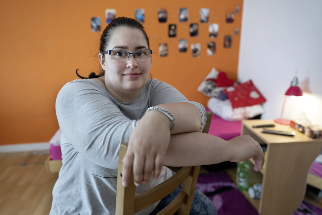 Eine Frau mit Brille sitzt entspannt verkehrt herum auf einem Stuhl in einem Raum mit einer orangefarbenen Wand. Im Hintergrund sind ein Bett, ein kleiner Schrank und eine Collage aus Fotos zu sehen. Sie tr&auml;gt ein graues Oberteil und lehnt sich bequem auf die Stuhllehne.